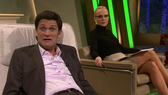 Der NDR-Moderator Christian Ehring liegt neben einer Frau auf einer Couch und macht einen erschrockenen Gesichtsausdruck.  