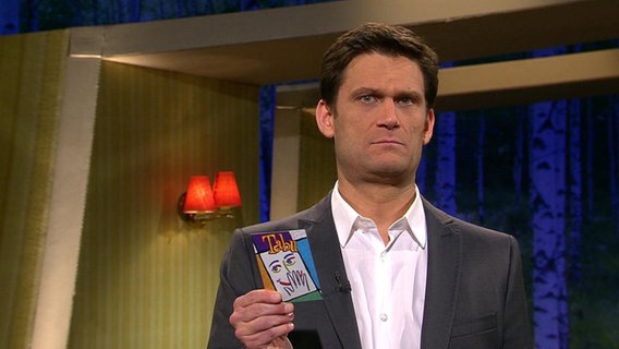 Der Moderator Christian Ehring hält eine Spielkarte in der Hand und macht ein fragendes Gesicht.  
