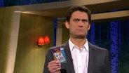 Der Moderator Christian Ehring hält eine Spielkarte in der Hand und macht ein fragendes Gesicht.  