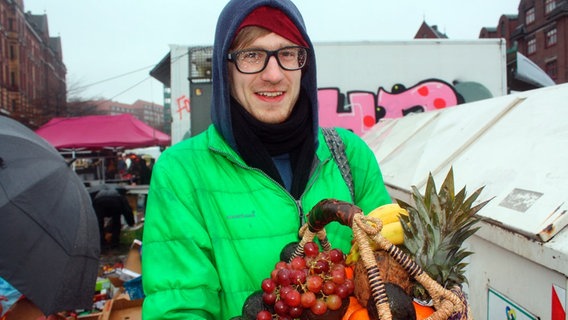 Student René steht auf dem Fischmark in Hamburg und hält einen mit Obst gefüllten Korb in die Kamera. © NDR/Ute Jurkovics 