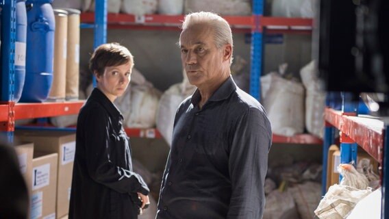 Eine Frau und ein Mann stehen in einer Halle zwischen Regalen mit Fässern, Kartons und Säcken. © NDR/BR/diwafilm GmbH 