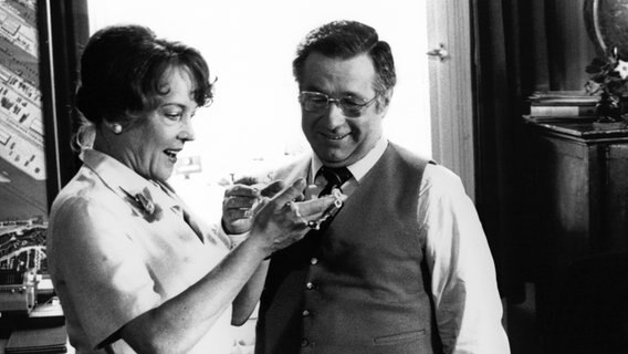 Makler Meier (Joachim Wolff) schenkt seiner Sekretärin Ilse (Hanna Seiffert) eine selbstgefertigte Kette aus Silberdraht. © NDR/Stroemer 
