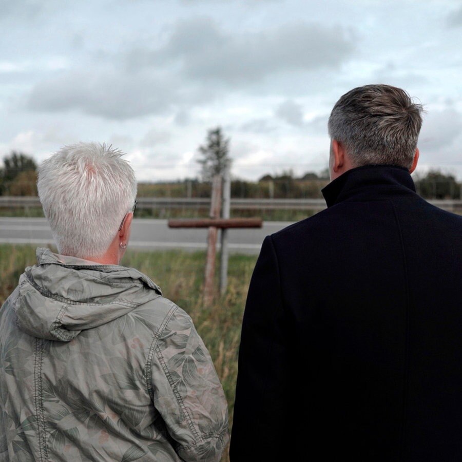 Eine Frau und ihr erwachsener Sohn stehen vor einem Holzkreuz an einer Straße. © NDR 