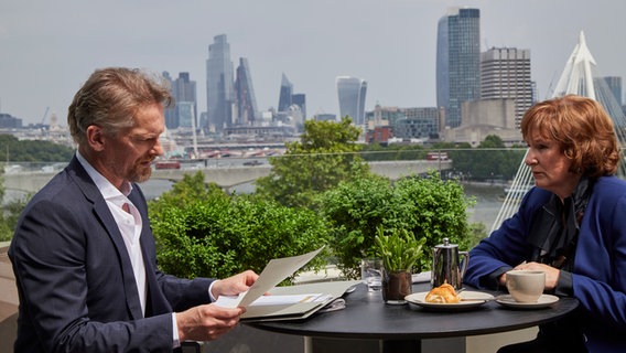 Szenenbild aus der Serie "The Split - Beziehungsstatus ungeklärt": Ein Mann und eine Frau sitzen an einem Cafe-Tisch mit Blick auf das Wasser und eine Skyline. © Sister Pictures Foto: Tereza Cervenova