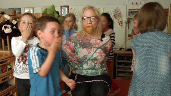 Mehrere Kinder tanzen um eine gefesselte Erzieherin.  