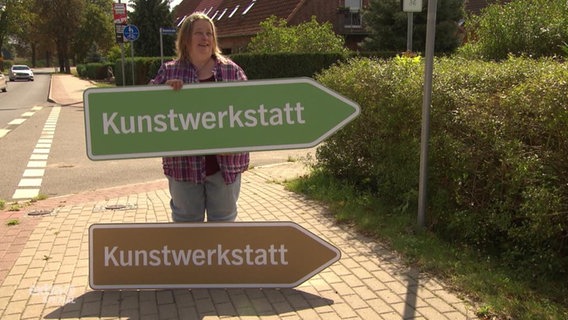 Frau hält zwei Wegweisschilder in die Höhe mit der Aufschrift "Kunstwerkstatt". Eins ist braun und eins ist grün.  