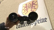 Ein Hand hält ein Fernrohr vor ein Schild mit dem Wappen des Landkreis Peine in die Luft  