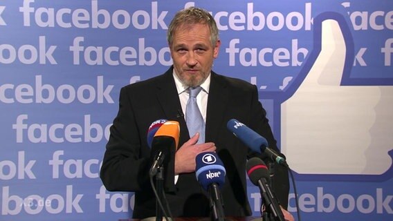 Ein Mann vor Mikrophonen, im Hintergrund das Facebook-Logo.  