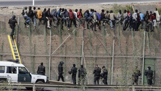 Flüchtlinge auf einer sehr hohen Mauer sitzend - am Boden Polizisten.  