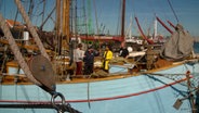 Vier Personen stehen auf einem Segelschiff in einem Hafen. © NDR 