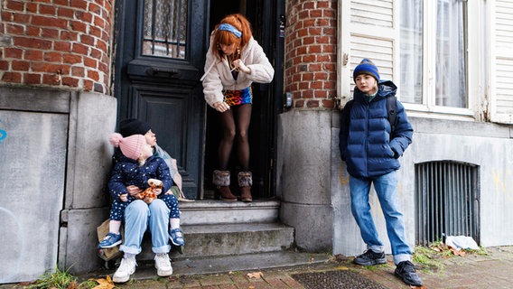 Szenenbild aus der französischen Serie "HIP: Ermittlerin mit Mords-IQ": Eine Frau blickt aus einem Hauseingang Kinder an. © NDR/Philippe Le Roux/TF1 Foto: Philippe Le Roux/TF1