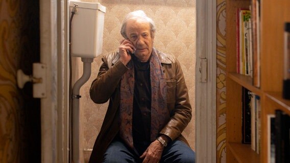 Szenenbild aus der französischen Serie "HIP: Ermittlerin mit Mords-IQ": Ein Mann sitzt auf einer Toilette und telefoniert. © NDR/Manuelle Toussaint Foto: Manuelle Toussaint