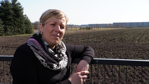 Eine Frau steht vor einem Feld mit einer großen Lagerhalle im Hintergrund. Eine Frau steht vor einem Feld mit einer großen Lagerhalle im Hintergrund. © Screenshot