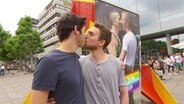 Ein schwules Pärchen küsst sich vor einem Plakat.  