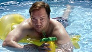 Charly Hübner liegt auf einer Luftmatratze im Pool und hält einen Drink in der Hand.  