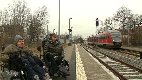 Zwei Rollstuhlfahrer warten am neuen Bahngleis, doch der Zug fährt am alten Gleis ein.  