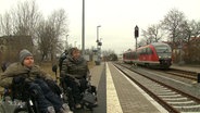 Zwei Rollstuhlfahrer warten am neuen Bahngleis, doch der Zug fährt am alten Gleis ein.  