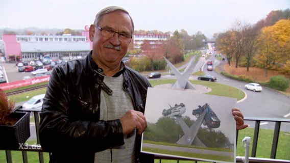 Ein Mann hält ein Bild in die Kamera auf dem Zwei Autos von einer Kunstinstallation aufgespießt wurden (Fotomontage)  