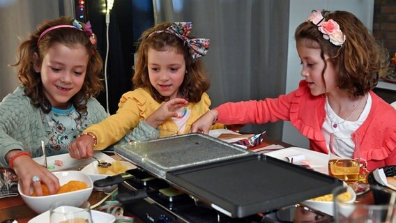 Mädchen-Drillinge sitzen am Tisch und essen Raclette. © NDR/Nico Maack 