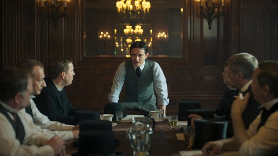 Franz Kafka (Joel Basman) steht am Kopfende eines Tisches und spricht zu sechs Männern, die rechts und links am Tisch sitzen. © Nicole Albiez 