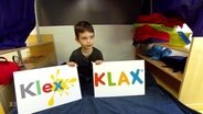 Ein Kind hält zwei Schilder, auf dem linken steht "Klex" und auf dem rechten ist "KLAX" zu lesen.  