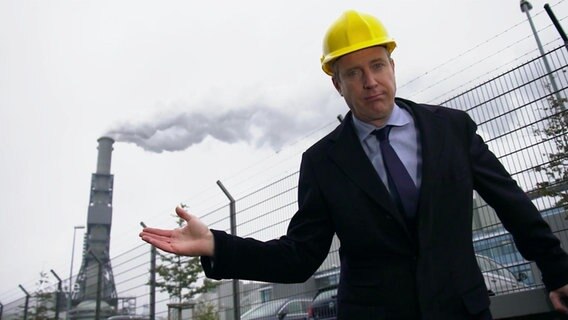 Ein Mann im Anzug mit einem Bauarbeiterhelm steht vor einem Kraftwerk.  