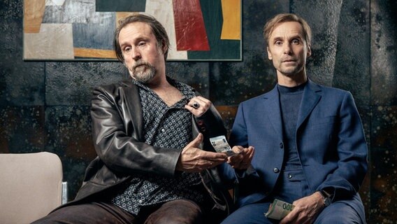 Szenenbild aus der Impro-Serie "Kranitz - Bei Trennung Geld zurück": Zwei Männer sitzen nebeneinander auf Stühlen und halten Geld in der Hand. © ARD Degeto/NDR/Thomas Leidig 