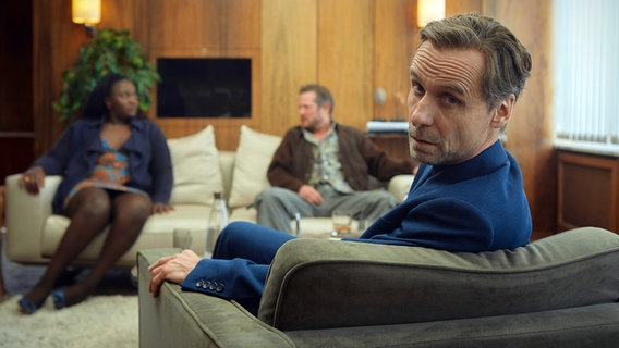Szenenbild aus der Impro-Serie "Kranitz - Bei Trennung Geld zurück": Ein Paar sitzt auf einem Sofa. Ein Mann auf einem Sessel blickt direkt in die Kamera. © ARD Degeto/NDR/Thomas Leidig 
