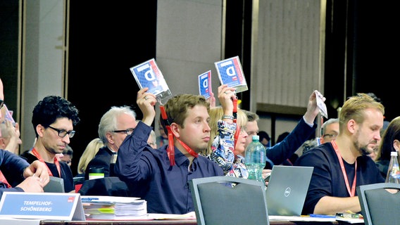 Kevin Kühnert sitzt in den Reihen anderer SPD-Mitglieder bei einer Veranstaltung und hält eine Wahlkarte hoch.  