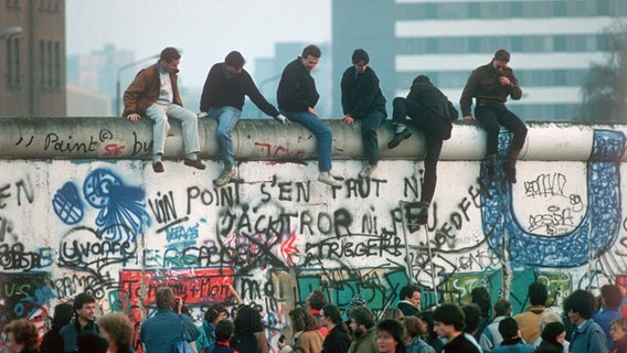 Sechs Männer sitzen auf der mit Graffiti bemalten Berliner Mauer. © picture alliance/dpa 
