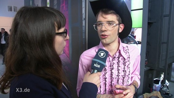 Eine Frau interviewt einen Mann, der ein rosa Hemd und einen Cowboyhut trägt.  