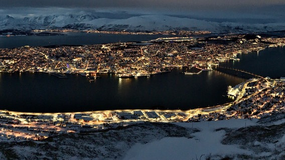 Eine beleuchtete Stadt liegt zwischen Wasser und vor schneebedeckten Bergen. © NDR Foto: Jörg Teiwes