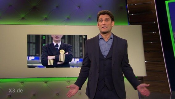 Der Moderator Christian Ehring, im Hintergrund ein Bild eines Piloten der die Arme verschränkt.  