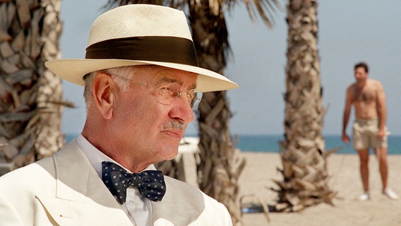 Ein Mann in einem weißen Anzug mit Fliege und Hut steht an einem Strand mit Palmen, im Hintergrund macht ein Mann Krafttraining. Ein Mann in einem weißen Anzug mit Fliege und Hut steht an einem Strand mit Palmen, im Hintergrund macht ein Mann Krafttraining. © WDR/Bavaria/Sibylle Anneck Foto: Sibylle Anneck