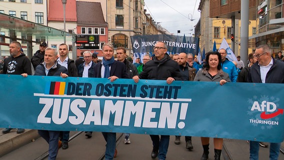 Eine Gruppe von Menschen marschiert auf einer Straße durch eine Stadt. Vorne wird ein Transparent mit einer Aufschrift gehalten. © WDR 