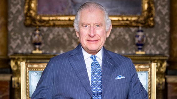 Der britische König Charles III. sitzt auf einem goldenen Stuhl und schaut lächelnd in die Kamera. © dpa Bildfunk 
