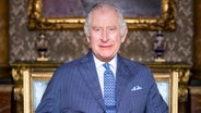 Der britische König Charles III. sitzt auf einem goldenen Stuhl und schaut lächelnd in die Kamera. © dpa Bildfunk 