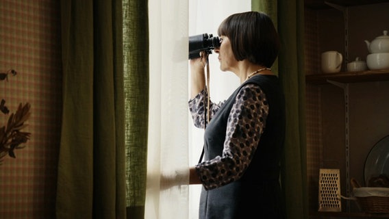 Eine Frau steht an einem Fenster und schaut durch ein Fernglas. © ARD Degeto/Conny Klein Foto: Conny Klein