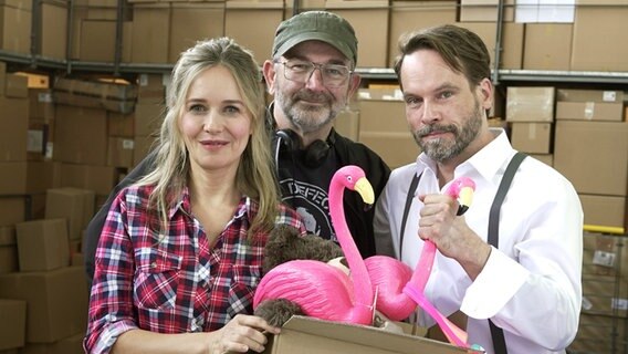 Bild zur NDR Serie "Retoure": Stefanie Stappenbeck, Torsten Wacker und Wanja Mues posieren mit Plastikflamingo für ein Drehstartfoto in einem Hochregallager. © NDR/Kinescope Film/Timo Schwarz 