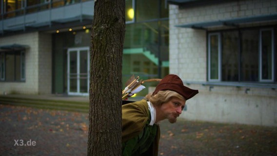 Ein als Robin Hood verkleideter Schauspieler schaut hinter einem Baum hervor.  