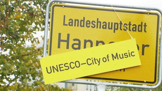 Ein Schild mit der Aufschrift UNESCO - City of Music hängt über Ortseingangsschilde der Landeshauptstadt Hannover  