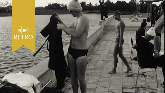 Schwimmer mit Bommelmütze zieht sich nach dem Wettkampf im Freibad schnell einen Bademantel über (1965).  