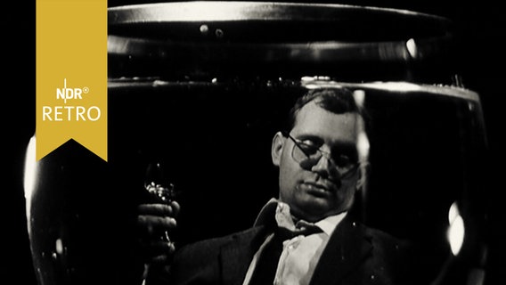 Wein trinkender Mann im Anzug in legerer Sitzpose spiegelt sich in einem Weinglas (1965)  