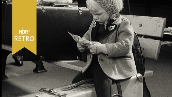 Kleines Kind mit Pudelmütze sittzt auf einem Koffer auf einem Bahnsteig und studiert einen Zettel (1965)  