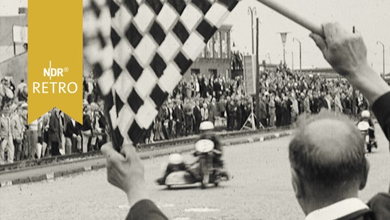 Motorradrennfahrer mit Sozius bei der Zieleinfahrt unter der Zielflagge des Schiedsrichters (1965)  