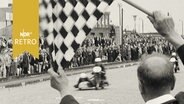 Motorradrennfahrer mit Sozius bei der Zieleinfahrt unter der Zielflagge des Schiedsrichters (1965)  