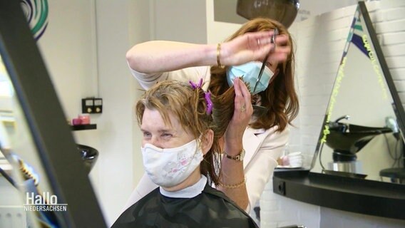 Eine Frisörin schneidet einer Person die Haare, beide tragen einen Mundschutz.  