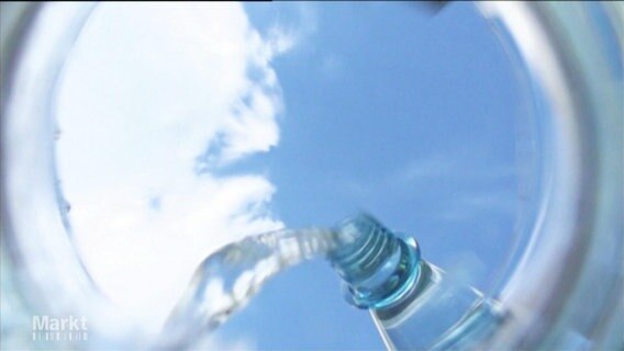 Sicht vom Boden eines Glases, in das aus einer Wasserflasche Wasser eingegossen wird. Darüber der blaue Himmel.  