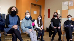 Schüler sitzen neben ihrer Lehrerin in einem Klassenraum. Alle tragen Masken. 
