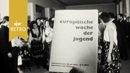 Programmheft "europäische woche der jugend" wird vor eine Gruppe von Jugendlichen in verschiedenen Trachten gehalten (1964)  
